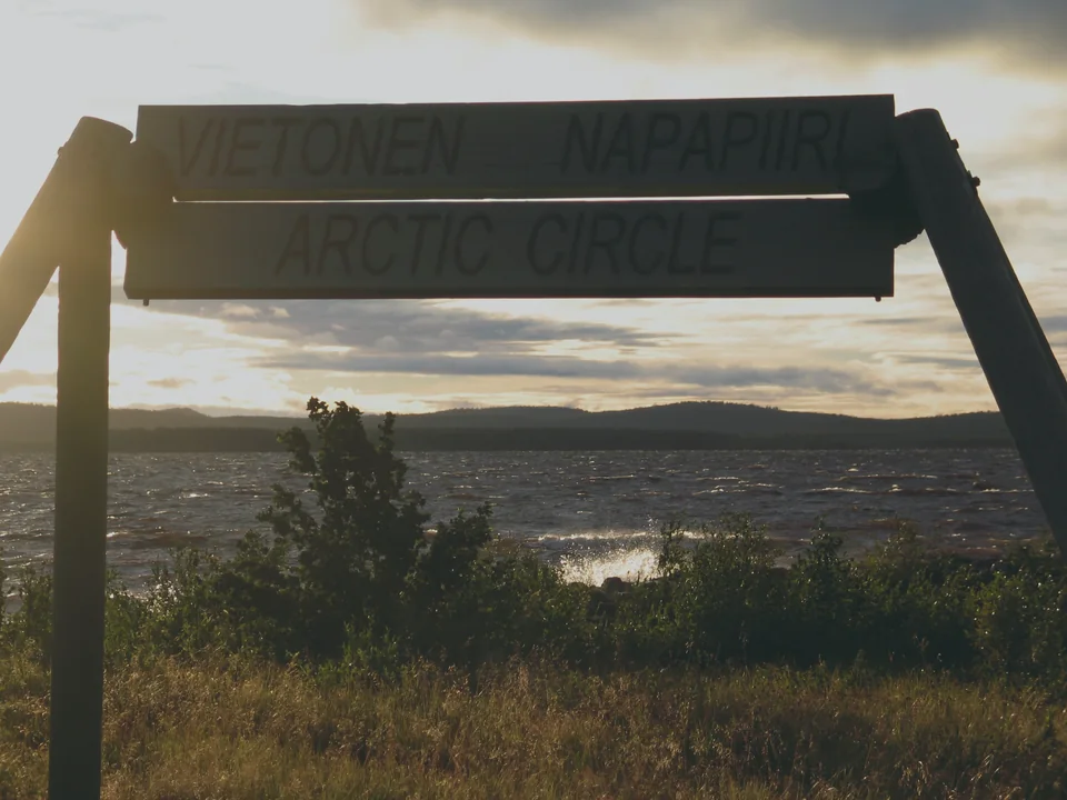 A wooden roadside sign reading Vietonen Napapiiri Arctic Circle, backlit by a low evening sun over a lake