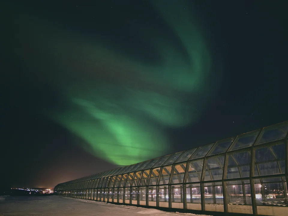 Arktikum's long glass-roofed corridor building at night, with a bright green aurora overhead