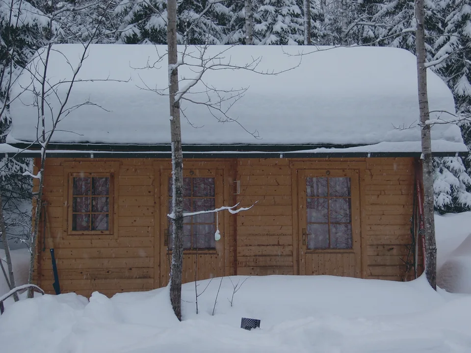 A timber cabin with a thick cap of snow on its roof, standing in deep snow between bare birches