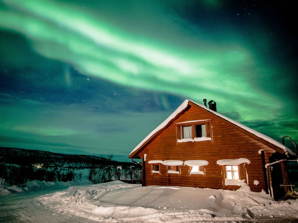 A wooden cabin with lit windows in deep snow near Kilpisjärvi, under a green aurora