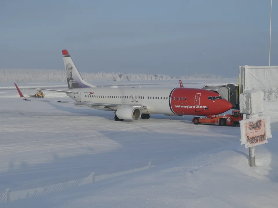 A red and white passenger jet parked at a snow-covered airport terminal under a pale winter sky, with frosted forest in the background