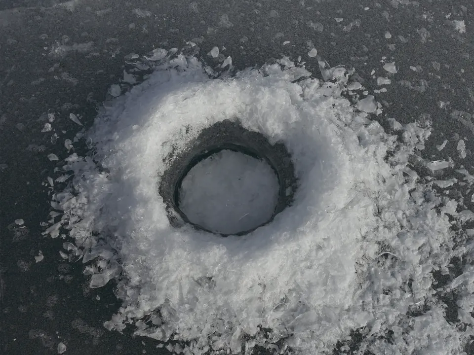 A freshly drilled ice-fishing hole in dark lake ice, ringed by a mound of white ice shavings