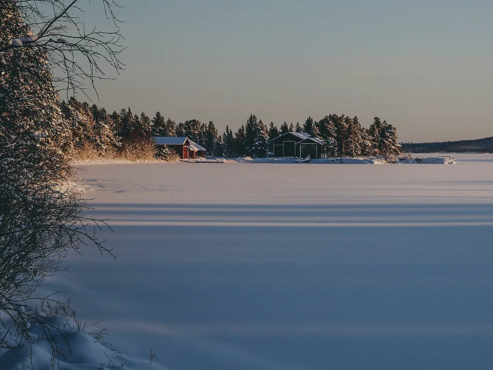 A frozen Lake Inari at low winter sun, with a red cabin and boathouse on the far shore and long blue shadows across the snow