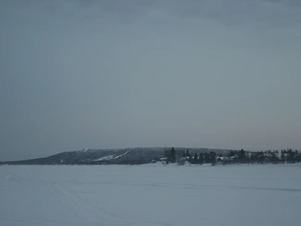 A wide frozen river at dusk under a grey-blue sky, with the forested Ounasvaara hill, its ski jump and a row of houses on the far bank