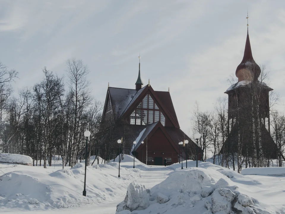 Kiruna's dark red wooden church and its separate bell tower standing in deep snow among bare birches under a pale sky