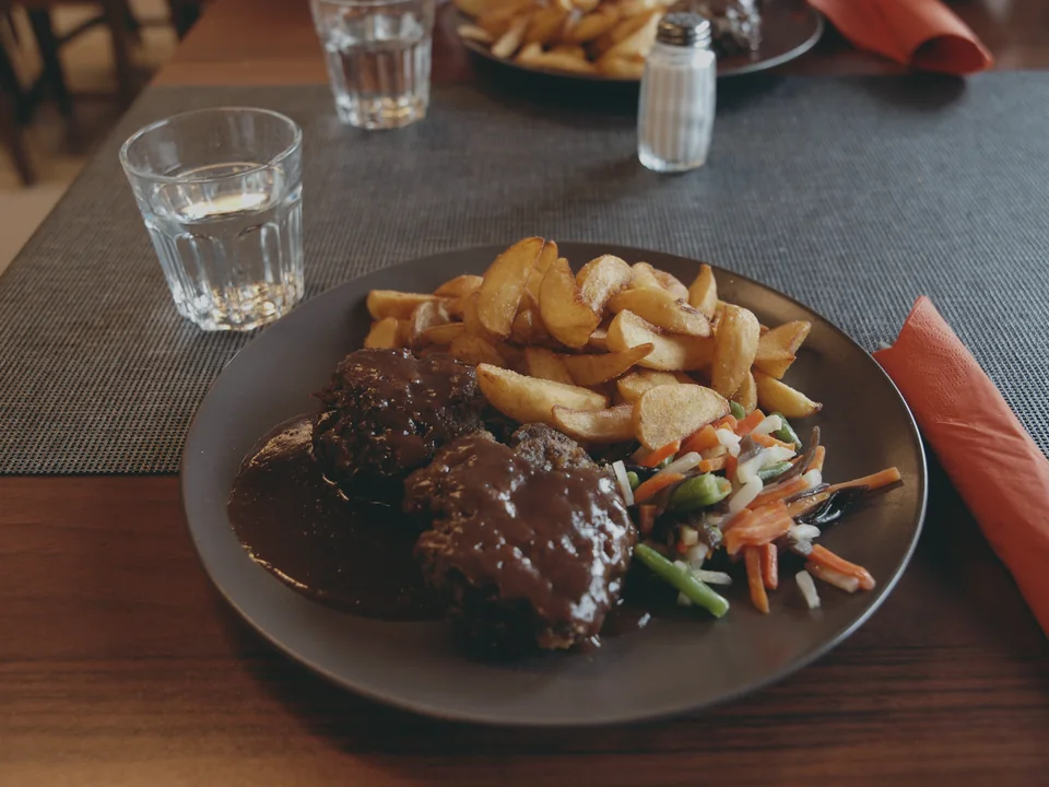 Two ground reindeer steaks in a dark gravy with potato wedges and mixed vegetables on a grey plate, a glass of water beside it on a restaurant table