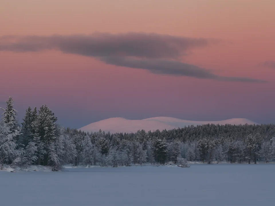 A snow-covered fell on the horizon behind a frozen lake and frosted forest, under a pink and blue dusk sky