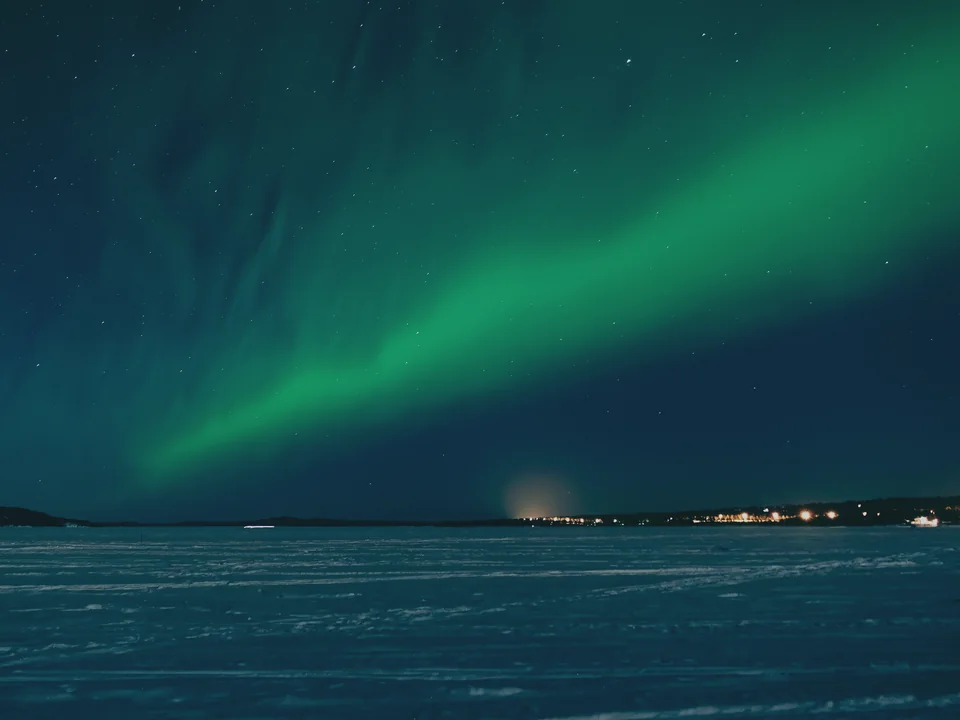 A green aurora band arcing across a deep teal night sky above a frozen lake, with the lights of a small settlement on the far shore