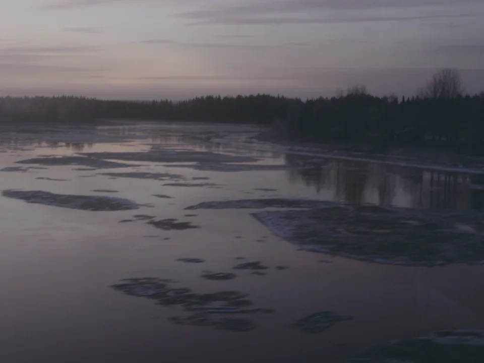 A wide river partly covered in early season ice, reflecting a pink and lilac dusk sky, with a frosted treeline on the far bank