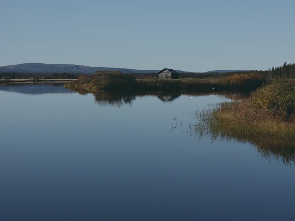A still lake reflecting a clear blue sky, with a wooden cabin and golden autumn grasses on the far shore and a fell on the horizon, at Suvanto village in Finnish Lapland