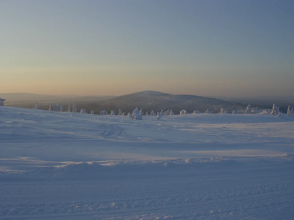 A snow-covered fell top at dusk, its silhouette against a pale orange horizon fading into blue sky, with frosted trees in the foreground and no people in view