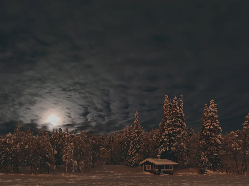 A snow-covered timber cabin glowing amber under moonlit, cloud-streaked winter trees at night in Sirkka, Levi's village, with no people in view