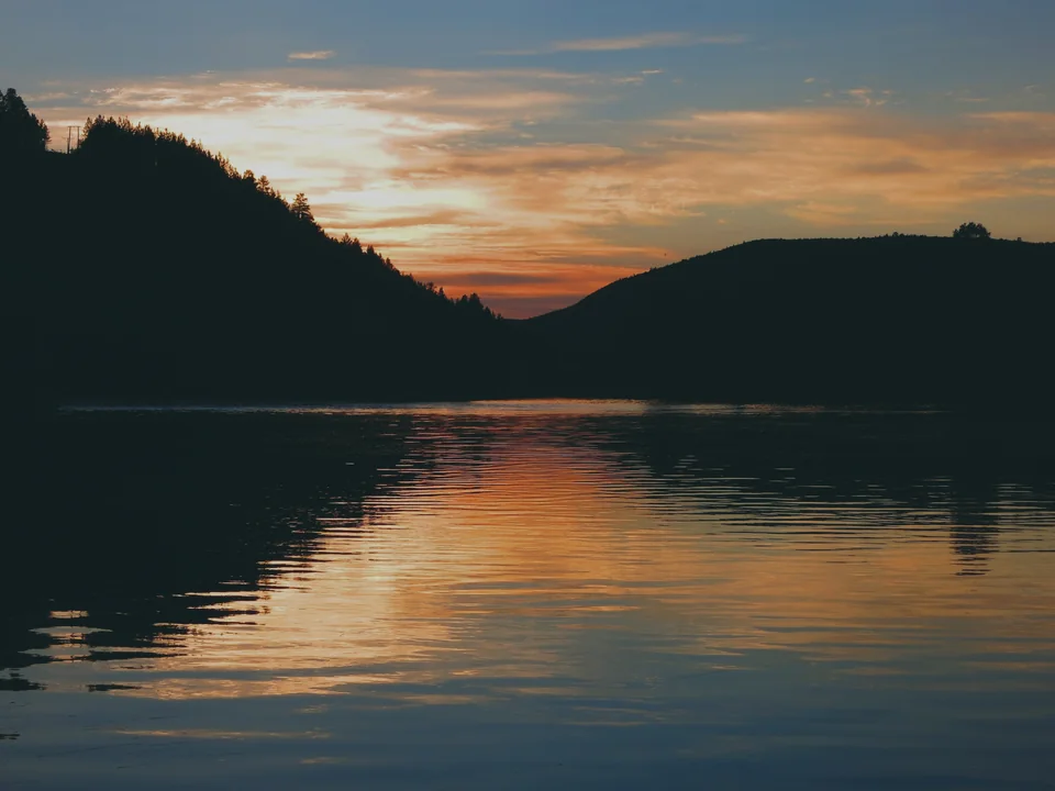A still northern lake at around half past midnight, the dark forested hills on either side in silhouette and an orange and gold sky reflected in the water