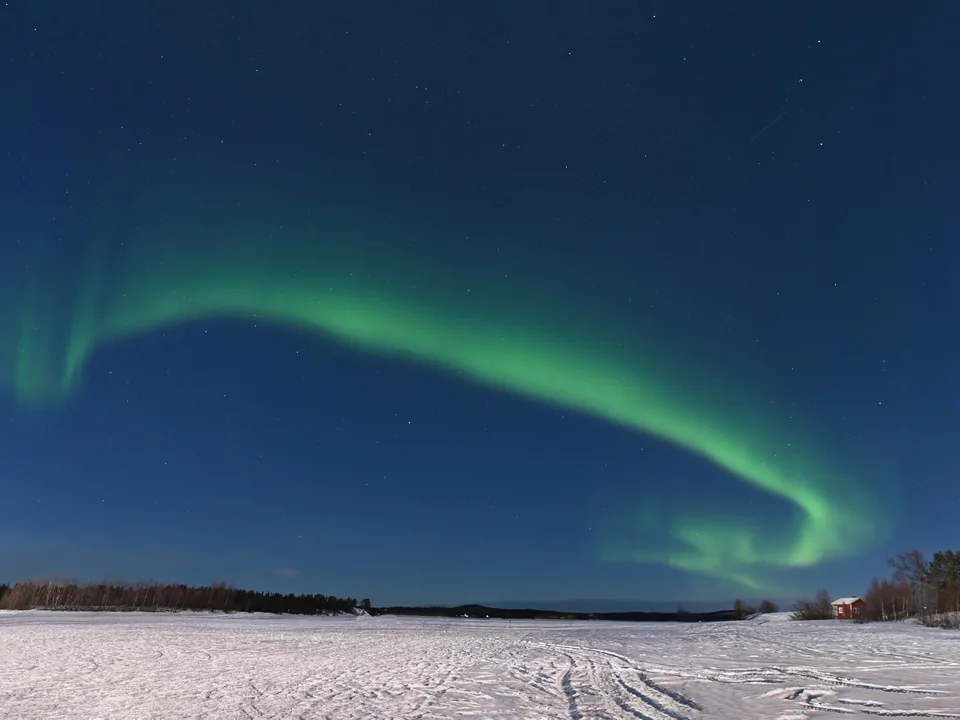 A green aurora arcing across a deep blue, moonlit sky over snow-covered open ground in Inari, in March