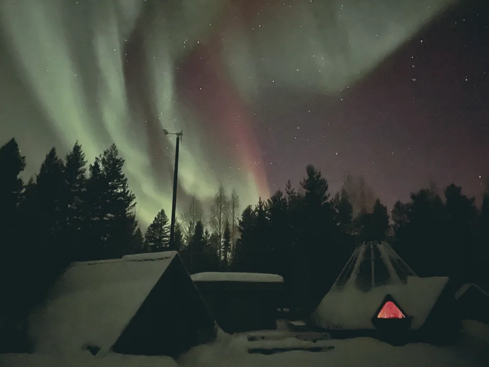 Green and red aurora over a snow-covered grillkota and log cabins in the Lapland countryside, with a small wind turbine and a warm light glowing inside the kota.