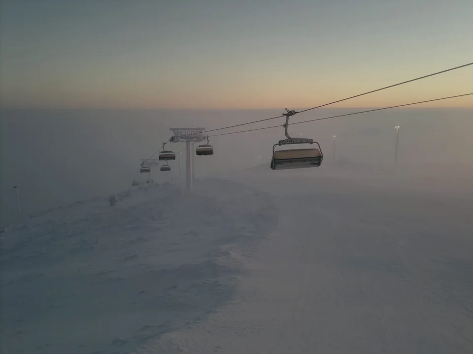 An empty chairlift climbing into thick winter mist at dusk, the sky fading from pale orange near the horizon to deep blue overhead, with snow-covered ground below and no people visible