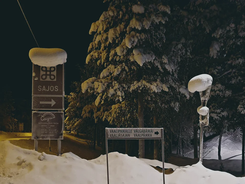 The Sajos sign lit at night beside snow-laden pine trees, with a directional sign for the building in the foreground