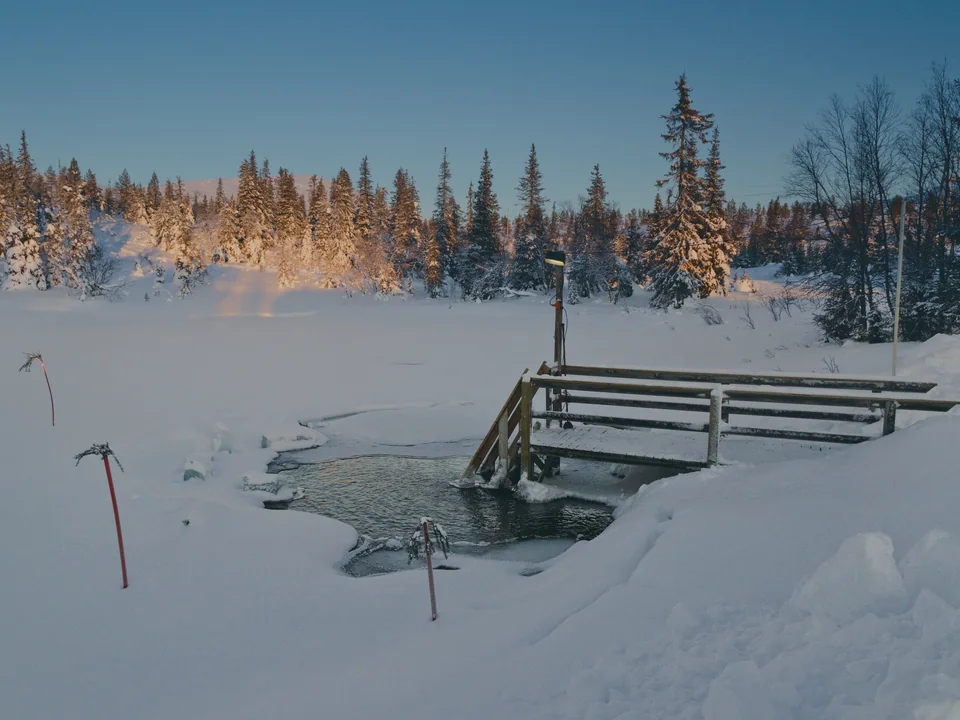 A swimming hole cut in the ice of a frozen pond, with a small wooden platform and ladder leading down to the dark water, deep snow all round and low sun lighting the snow-laden spruces behind, with nobody in view