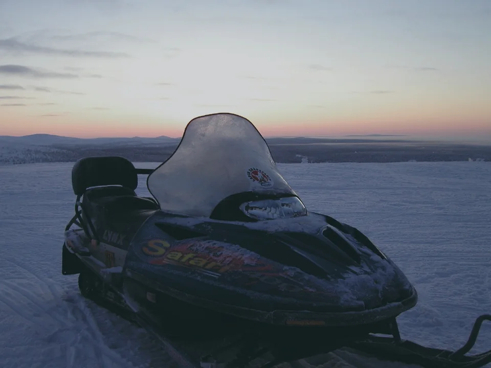 A single parked snowmobile, frosted over, on top of a fell in Saariselkä at dusk, with a wide snow-covered landscape and a pink and blue sky behind it, nobody in view