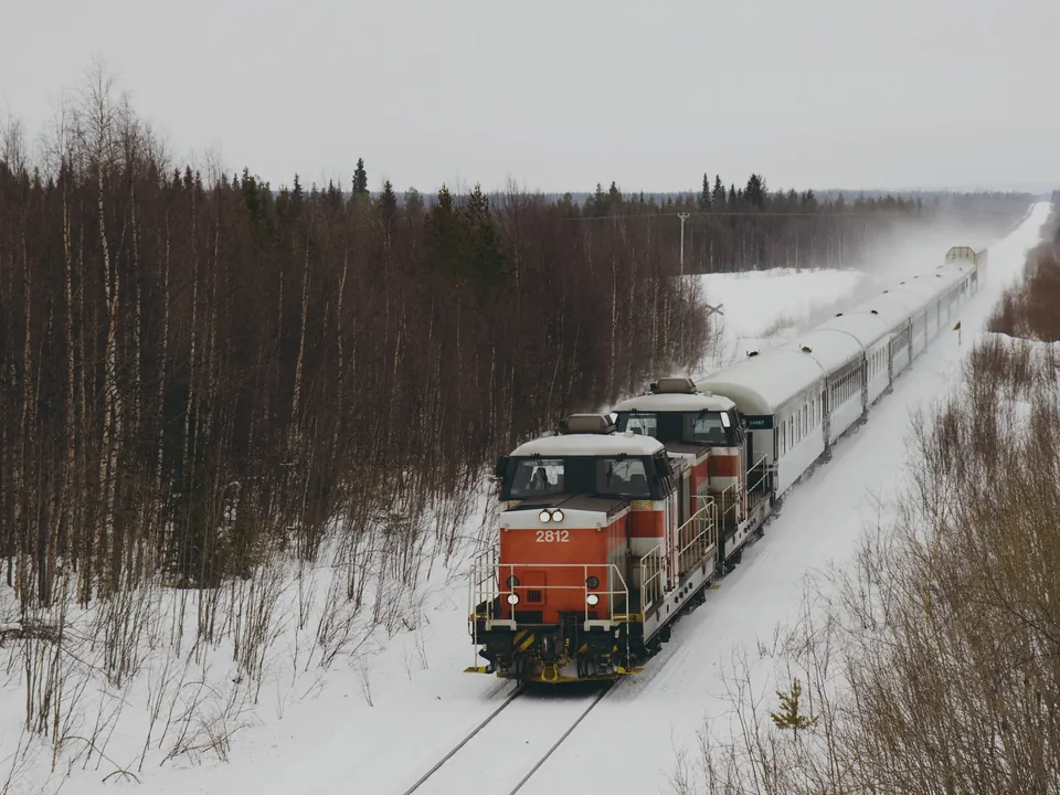 A VR night train, hauled by two orange and white diesel locomotives, crossing a snow-covered clearing lined with birch and spruce near Kolari.