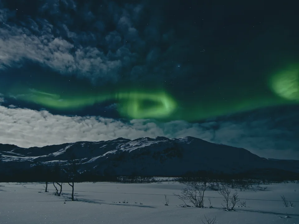 Green aurora coiling above a snow-covered mountain ridge, with a bank of cloud across the lower sky and bare scrub on the snowfield in front