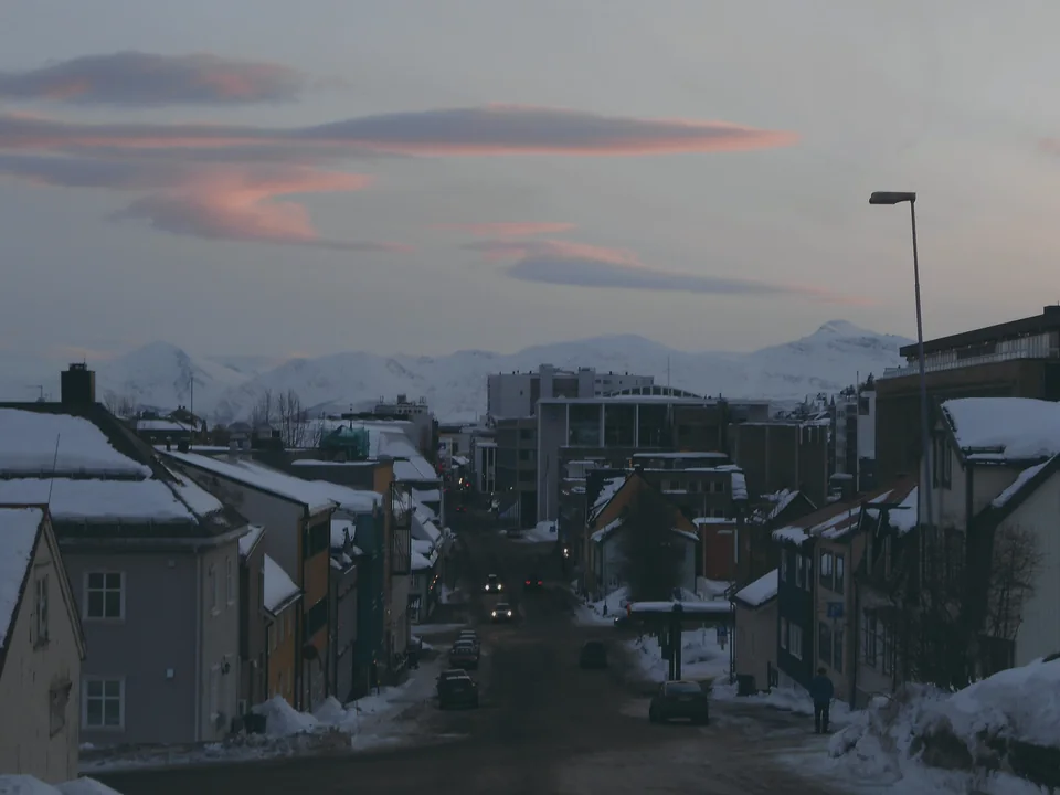 A snowy street of painted wooden houses running downhill towards the fjord, with pink cloud over the mountains beyond