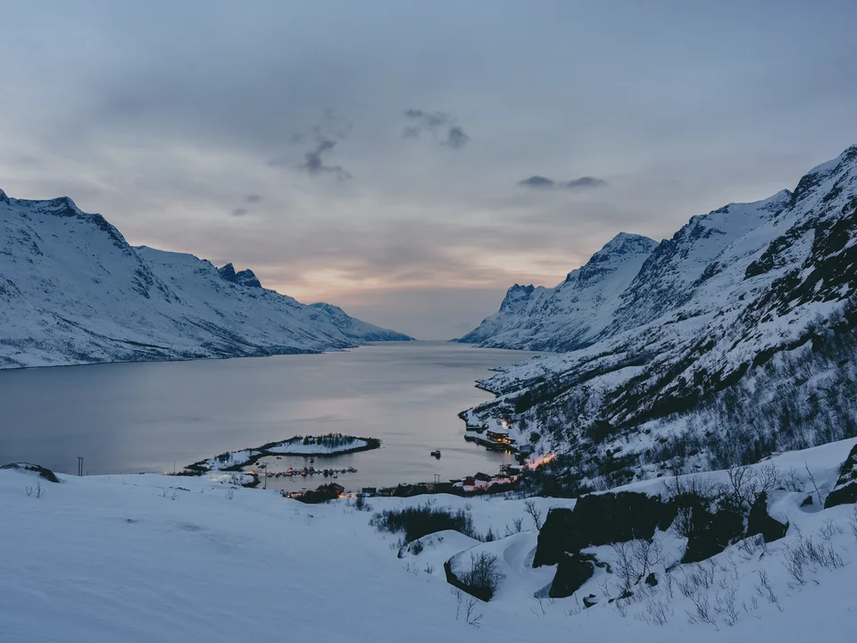 A fjord running between steep snow-covered peaks after sunset, the water still, a few small buildings on the far shore under a pink and grey sky