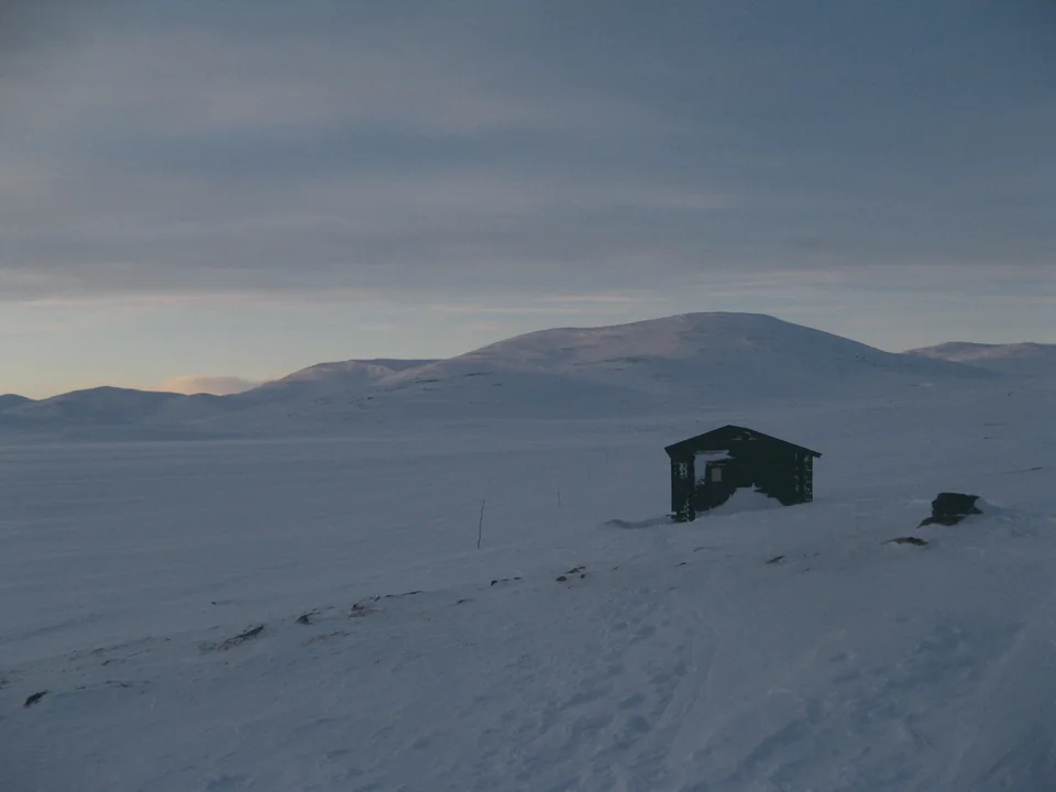 The Pihtsusjärvi wilderness hut in deep snow below rounded fells in Enontekiö, in the evening light of March