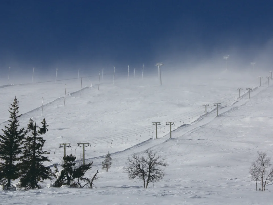 A ski slope climbing into blowing snow and mist under a deep blue sky, chairlift towers marching up the fellside, with frosted spruce trees in the foreground and nobody in the frame