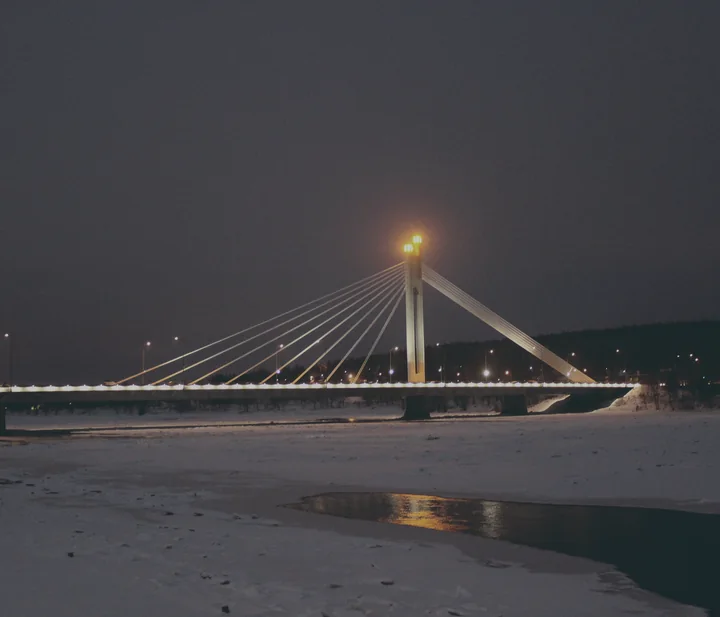 The Lumberjack’s Candle Bridge in Rovaniemi after dark, its pylon lit, over a river edged with snow and ice