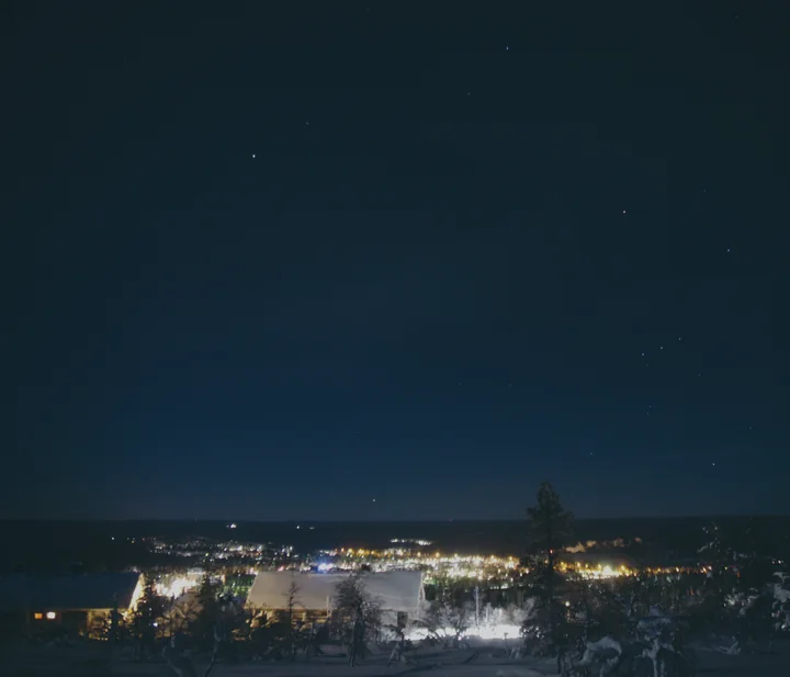 Saariselkä at night from the top of Kaunispää fell, snow-covered roofs in front and the village lights in the valley below a starry sky