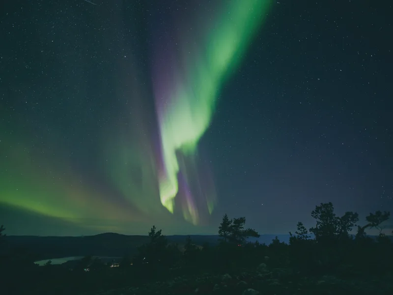 An aurora curtain in green and violet over Levi fell in September, low pines on the fell top and a lake in the valley below